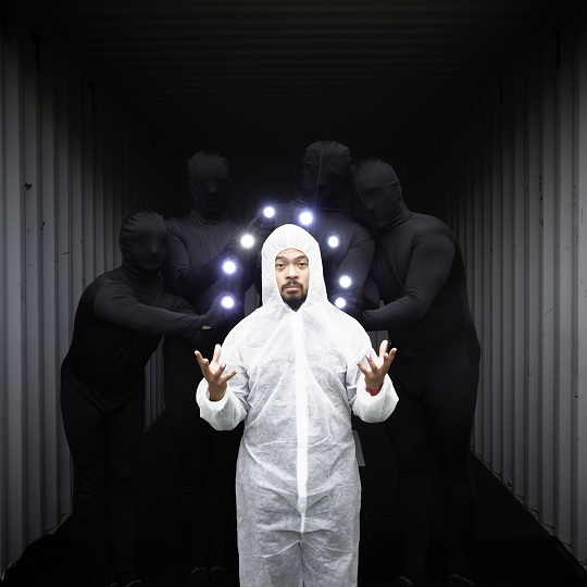 Photo of person in a white hazmat type body suit with hood, the person has a black goatee and has their hands mid air  with fingers pointing up, they hold glowing lit up type balls that make an arc shape abover their hands, in the background is a group of four performers in black bodysuits that also cover their faces they stand in a grey shipping container (you can only see the corrugated sides of the container